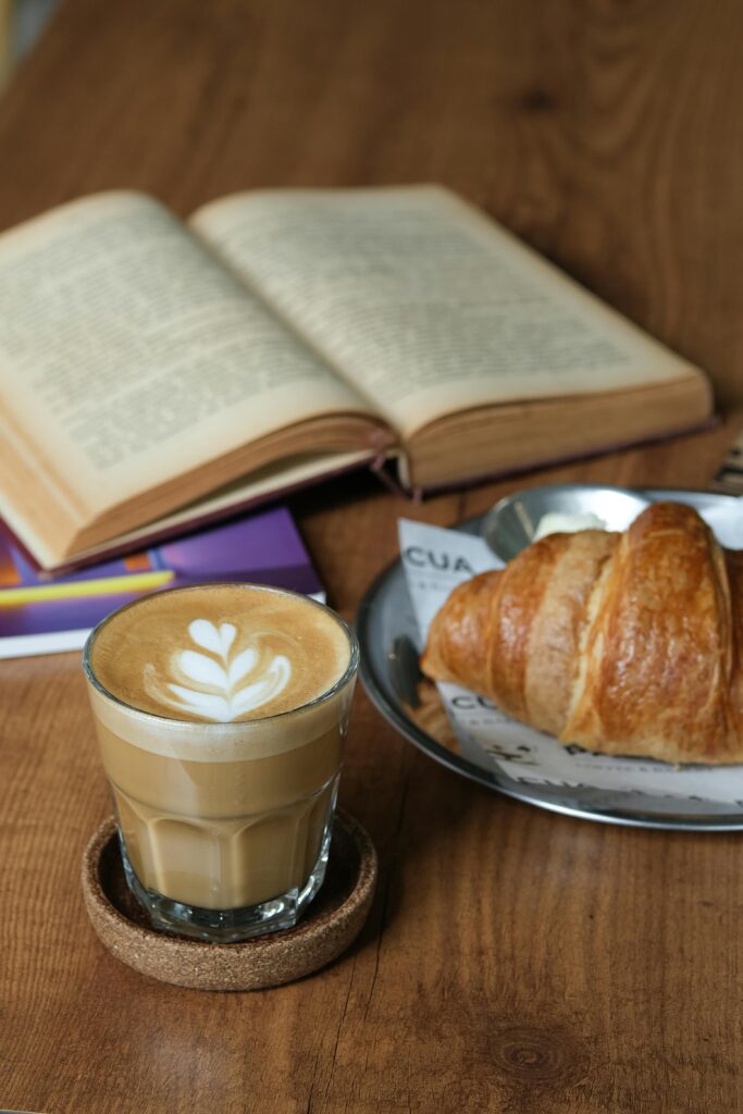 Warm and inviting setup of a latte, croissant, and open book on a wooden table.
